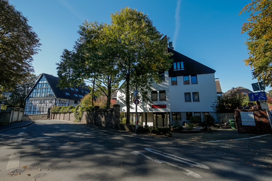 Ein Blick auf die Straßenkreuzung vor dem Gästehaus. Das Gästehaus ist im Hintergrund zu erkennen und wird teilweise von Bäumen verdeckt.