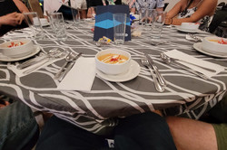 Hier sieht man ein Tisch, an dem mehrere Menschen sitzen, und an den gerade eine Suppe serviert worden ist. 