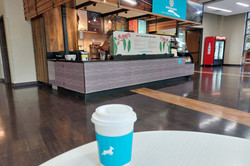 Kleines Cafe an der Uni Hier sieht man einen To Go Kaffeebecher, der auf einem Tisch steht. Im Hintergrund sieht man eine Theke.