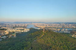 Hier sieht man Seoul von oben. Man sieht zunächst viel Grünfläche, die dann in eine Stadt übergeht.