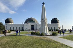 Ein weißes Gebäude mit schwarzen Kuppeldächern und einer weißen Säule im Vordergund. Drumherum ist viel grüne Wiese und einige Menschen stehen vor dem Observatorium. 