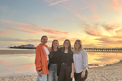 Vier Mädchen stehen Arm in Arm am Strand und lächeln in die Kamera. Im Hintergrund ist das Meer zu sehen und der Himmel ist von der untergehenden Sonne leicht rosa verfärbt. 