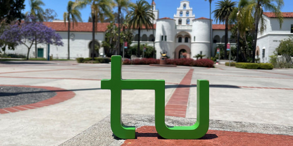 ein TU Logo steht auf dem Campus der SDSU vor einem weißen Gebäude im mexikanischen Stil. Palmen sind auf beiden Seiten vom Gebäude