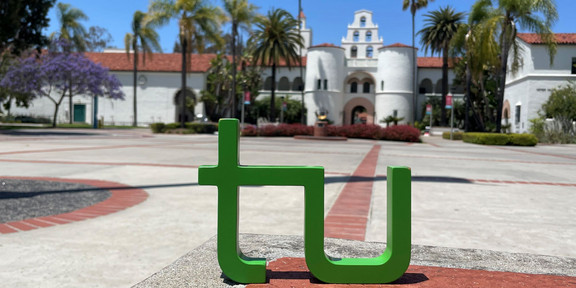 ein TU Logo steht auf dem Campus der SDSU vor einem weißen Gebäude im mexikanischen Stil. Palmen sind auf beiden Seiten vom Gebäude