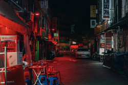 Das erste Bild Blick in eine schmale Straße bei Nacht, die mit rotem Licht beleuchtet ist. Auf dem Boden ist ein großes X aufgemalt und links sind einige geöffnete Shops zu sehen.