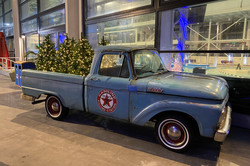 Ein blauer Oldtimer-Pick-up in einer großen Halle. Auf der Ladefläche liegen Weihnachsbäume, die mit Lichterketten geschmückt sind. 