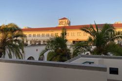 Hier sieht man ein langes Gebäude im spanischen Baustil, welches von der Abendsonne angestrahlt wird. Im Vordergrund stehen einige Palmen. 