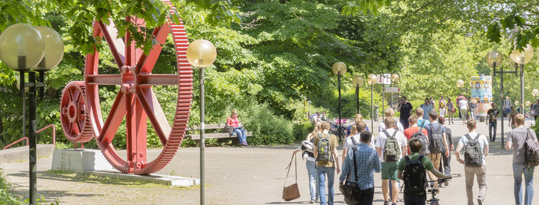 Studierende laufen über den Campus. Es ist eine große rote Skulptur in Form von großen Zahnrädern zu erkennen.