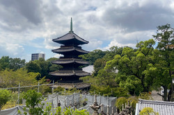 Hier sieht man einen kleinen Tempel, umgeben von vielen Bäumen und im Hintergrund ragt ein Hochhaus empor.  