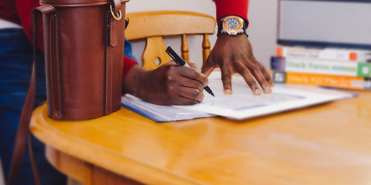 Ein Mann unterschreibt ein Dokument, das auf einem Tisch liegt. Neben dem Dokument liegt eine braune Ledertasche. 