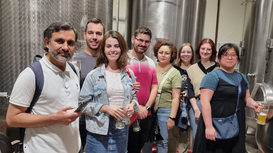 Eine Gruppe von Menschen steht vor den großen Metallfässern einer Brauerei. 