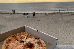 Ein Karton mit Pizza und im Hintergrund ein Strand mit dem Meer bei Sonnenuntergang. 