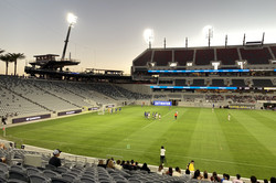 Ein Fußballstadion in der Dämmerung mit ein paar Spielerinnen auf dem Feld. Die Tribüne ist fast leer und die Scheinwerfer beleuten das Spielfeld. 