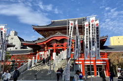 Ein großer Tempel, zu dem eine breite Treppe hinaufführt und vor dem viele weiße Fahnen wehen, auf denen in schwarzer Farbe japanische Schriftzeichen geschrieben stehen. Ringsherum sind Plattenbauten zu erkennen und der Himmel ist strahlend blau. 