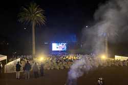 Es zeigt ein Outdoor Kino, gant vorne ist die Leinwand und davor liegen viele Sitzkissen, hinter den Plätzen sind kleine Fackeln aufgestellt.