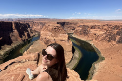 Horseshoe Bend Storytellerin Lisa sitzt im Vordergrund auf einem roten Felsen. Im Hintergrund verläuft ein Fluss durch eine Felsschlucht in Hufeisenform.