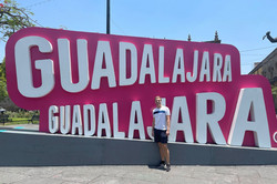 Hier sieht man den Studenten vor einem pink/weißen Schild, auf dem Guadalajara steht. 