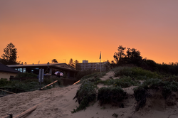 Es zeigt einen Sonnenuntergang an einem Sandstrand, im Hintergrund stehen Häuser und Bäume.