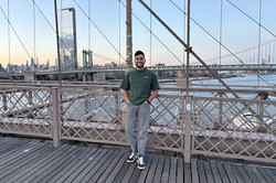 Es zeigt Aykut auf einer Brücke, im Hintergrund ist Wasser und die Skyline von New York 