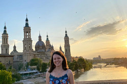 Hier sieht man die Studentin im Sommer auf einer Brücke über einem Fluss stehen mit einem Schloss im Hintergrund.