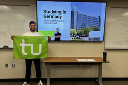 Aykut steht mit einer Flagge mit dem TU Logo vor einer Leinwand. Auf der Leinwand wird eine Präsentation mit dem Titel Studying in Germany gezeigt.