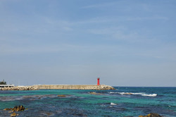 Hier sieht man das Meer mit einem langen Felssteg, an dessen Ende ein roter Leuchtturm steht.