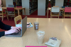 Hier sieht man einen pinken Notizblock auf dem Tisch. Am Rand sieht man eine Studentin am Laptop. 