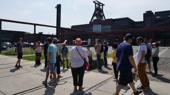 Menschen stehen zuhörend auf dem Gelände der Zeche Zollverein mit dem Förderturm im Hintergrund.
