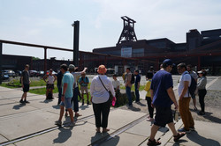 People are standing on the premises of Zeche Zollverein and listening, with the shaft tower in the background.