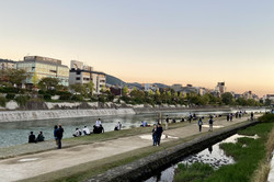 Entspannte Abendstimmung am Kamogawa-Fluss Aussicht auf einen Fluss, an dessen Ufer einige Menschen sitzen oder spazieren. Auf der anderen Seite des Flusses stehen einige Gebäude. Der Himmel ist vom Sonnenuntergang leicht rötlich gefärbt.