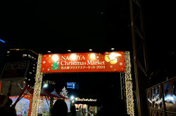 Weihnachtsmarkt in Nagoya Ein beleuchtetes Schild mit der Aufschrift "Nagoy Christmas Market" und einige weihnachtlich leuchtende Stände im Hintergrund in der abendlichen Dunkelheit.