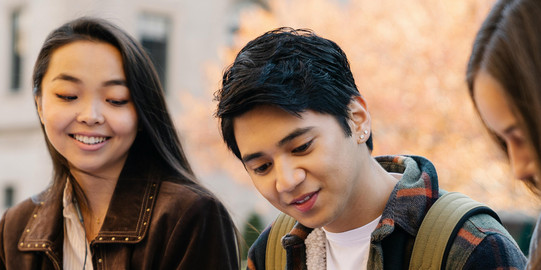 Two young women and a young man are sitting on a bench, sharing scripts and smiling in front of a university building with autumn trees.