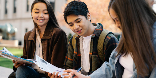 Two young women and a young man are sitting on a bench, sharing scripts and smiling in front of a university building with autumn trees.