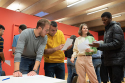 Some international students are standing next to a table and talking. They are showing each other on pieces of paper.