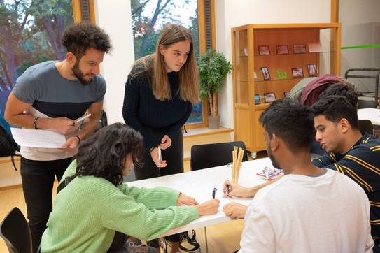 Some international students are sitting and standing around a table. They are talking to each other.