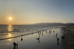 Sonnenuntergang über dem Meer mit weitläufigem Strand und vielen Menschen im und am Wasser. Im Hintergrund sind Berge zu sehen. 