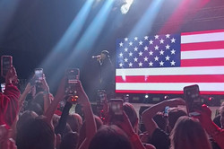 Greenfest Konzert von YG Man sieht einen dunklen Raum mit einer Bühne, vielen Lichtern und Menschen und einer USA Flagge.