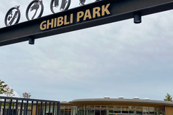 Das Eingangstor mit der Aufschrift "Ghibli Park", dahinter ein großer gepflastereter Platz und ein rundes Gabäude mit Glasfassaden. 