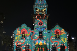 Es zeigt ein Gebäude bei Nacht, es wird bunt beleuchtet und an der Fassade sind viele bunte Symbole, wie Schmetterlinge abgebildet.