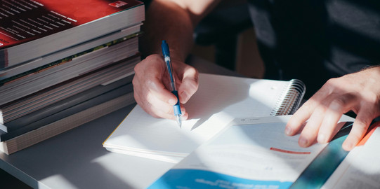 Person is writing; books are on the table; there are light and dark accents in the picture