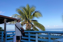 Veranda am Strand Es zeigt Will seitlich, mit einer Gitarre in der Hand. Er steht auf der Veranda und davor steht eine große Palme, im Hintergrund ist das Meer.