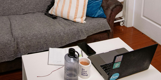 Ein gemütliches Wohnzimmer mit einem grauen Sofa, bunten Kissen und einem weißen Tisch. Auf dem Tisch stehen eine Wasserflasche, eine Tasse Kaffee und ein Laptop. An der Wand hängen Fotos und Bücherregale.