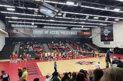Ein Volleyballfeld mit einigen Spielerinnen und Zuschauern auf der Tribüne hinter dem Spielfeld. An der Wand steht "Home of Aztec Volleyball" geschrieben. 