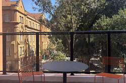 Blick auf einen großen Balkon mit einem Bistrotisch und ein paar Stühlen. Im Hintergrund stehen ein altes Gebäude und viele Bäume