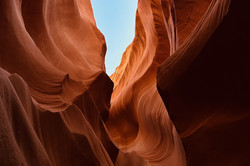 Rote, geschwungene Felswände bilden rechts und links eine Schlucht. Im Hintergrund strahlt der blaue Himmel durch den Canyon. 