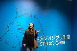 Die Studentin vor einer blauen Wand, auf der mit schwarzer Farbe eine Kreatur gezeichnet ist. Rechts steht "Studio Ghibli" geschrieben. 