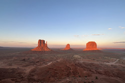 Monument Valley Felslandschaft mit drei großen, roten Felsen vor blauem Himmel, die von der untergehenden Sonne angeleuchtet werden.