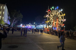 Hier sieht man ein bunt beleuchtetes Riesenrad in der Nacht mit vielen Menschen davor. Links daneben befinden sich weitere beleuchtete Fahrgeschäfte. 