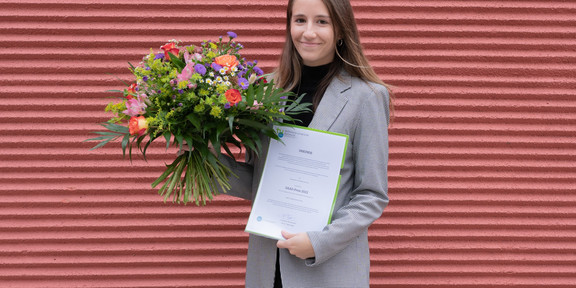 A young woman in a blazer stands in front of a brick wall holding a bouquet of flowers and a certificate.