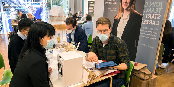Two men are sitting at a table in front of a Miele display. One of the men is showing something on his tablet to a woman sitting opposite him. Between them is a miniature washing machine. Other information stands can be seen in the background. 
